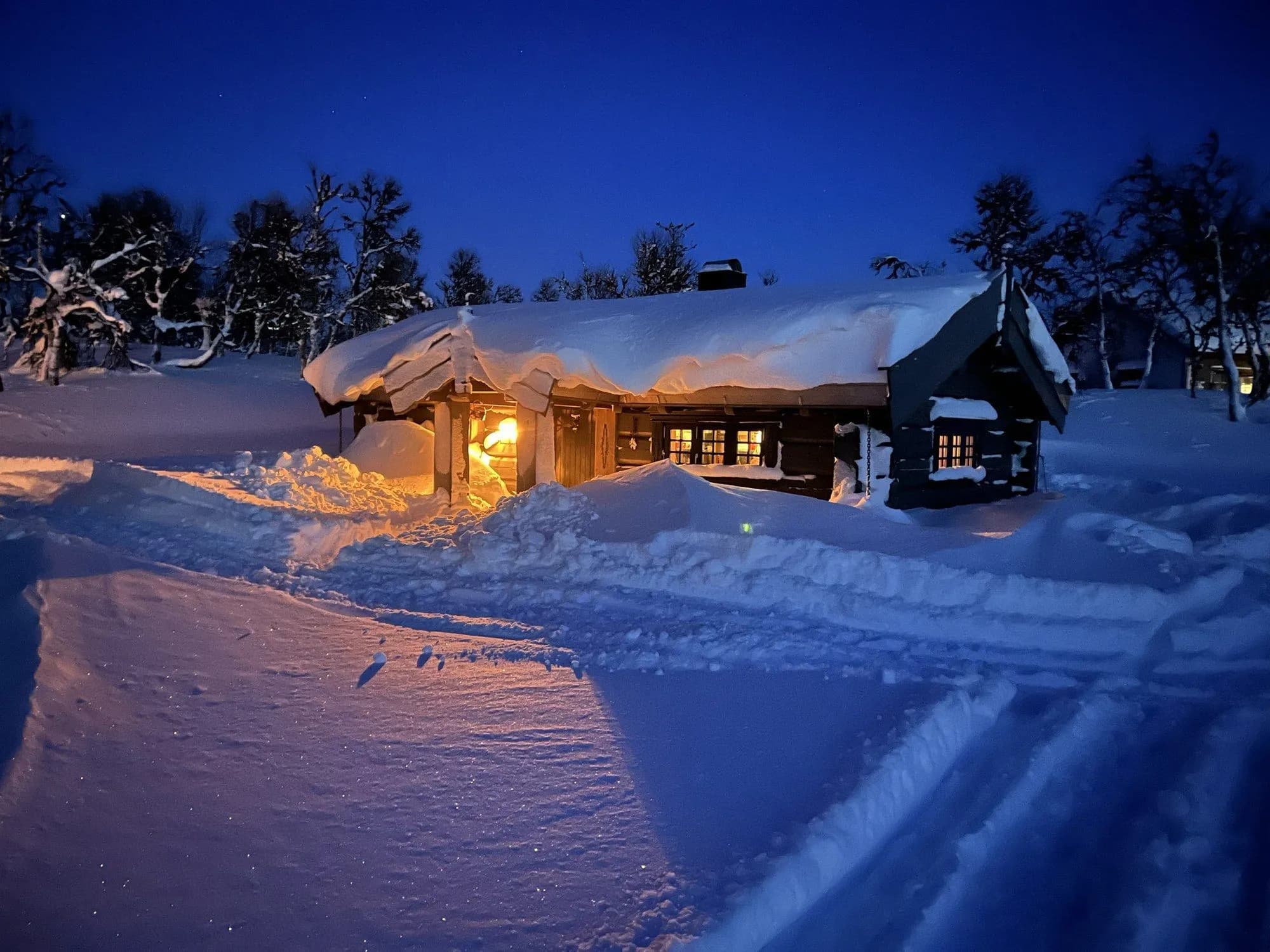 Anneksset dypt i snø en vinterkveld, med varmt lys fra vinduene i blåtimen.
