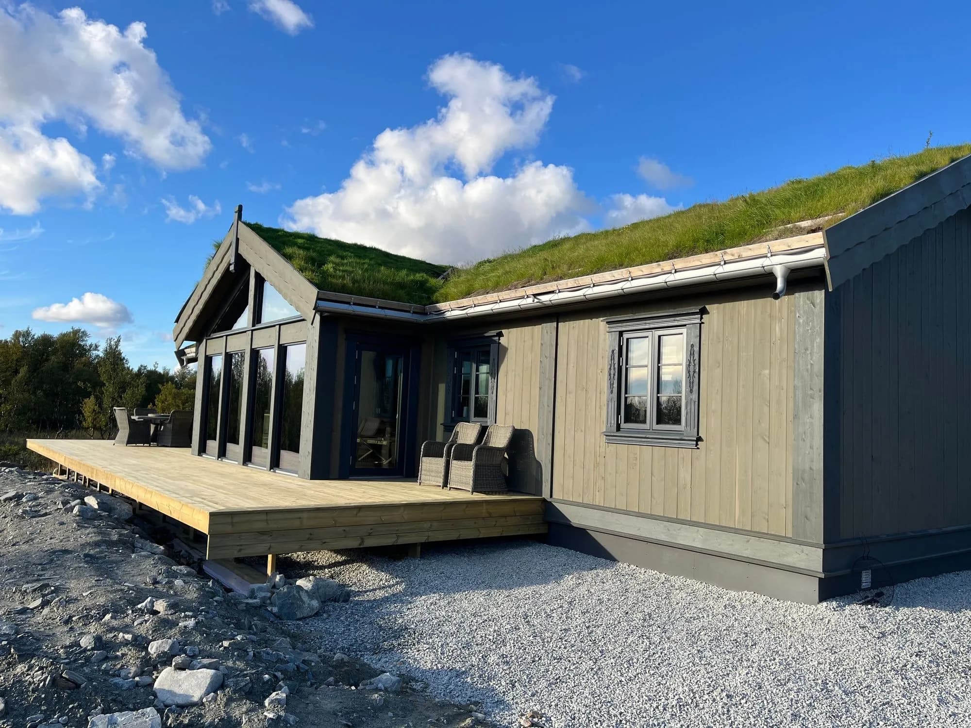 Moderne fjellhytte med torvtak og store vinduer, sett fra terrassesiden en solrik sommerdag.