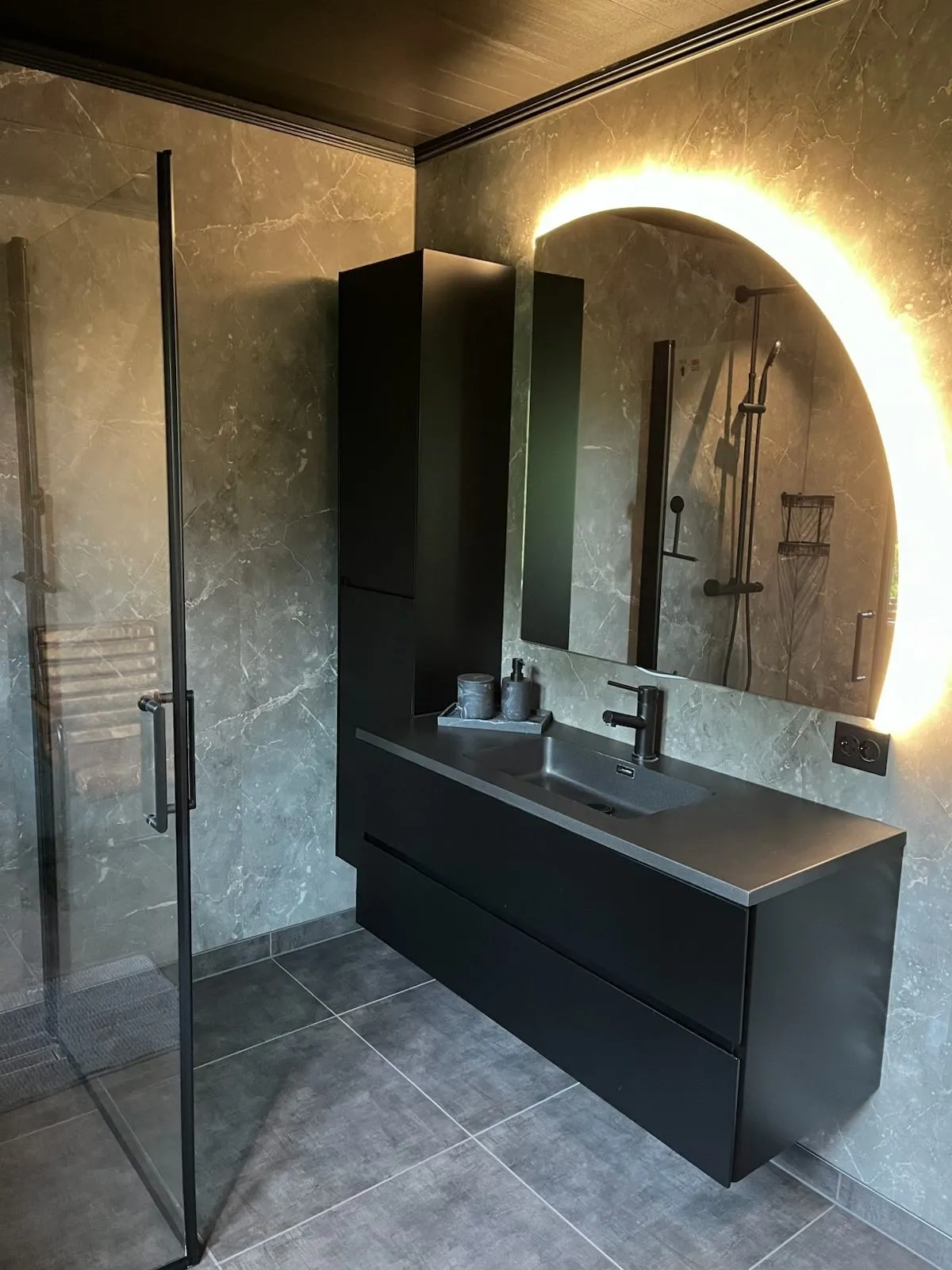 Det store badet sett mot servanten — mørk vegghengt servantmøbel med grå benkeplate, rundt LED-opplyst speil og innglasset dusj til venstre.