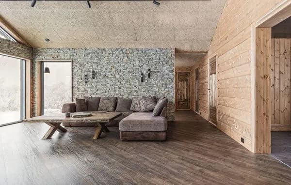 Stue med natursteinvegg, stor stoffsofa i hjørne, rustikt plankebord og store vinduer mot snølandskapet.