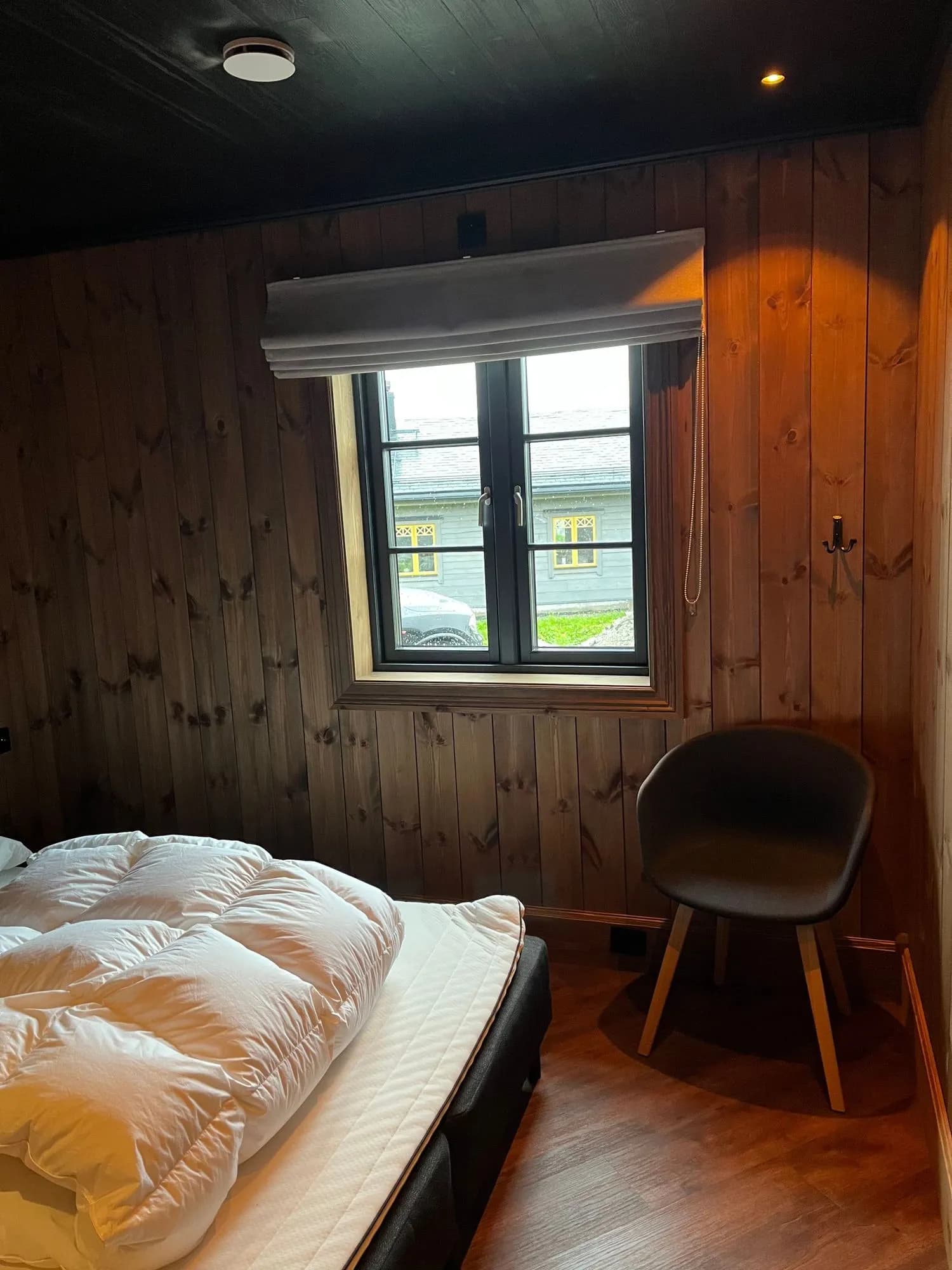 Soverom 1 med oppredd dobbeltseng, mørkbeisede furupanel-vegger og et vindu med rullegardin ut mot nabohyttene.